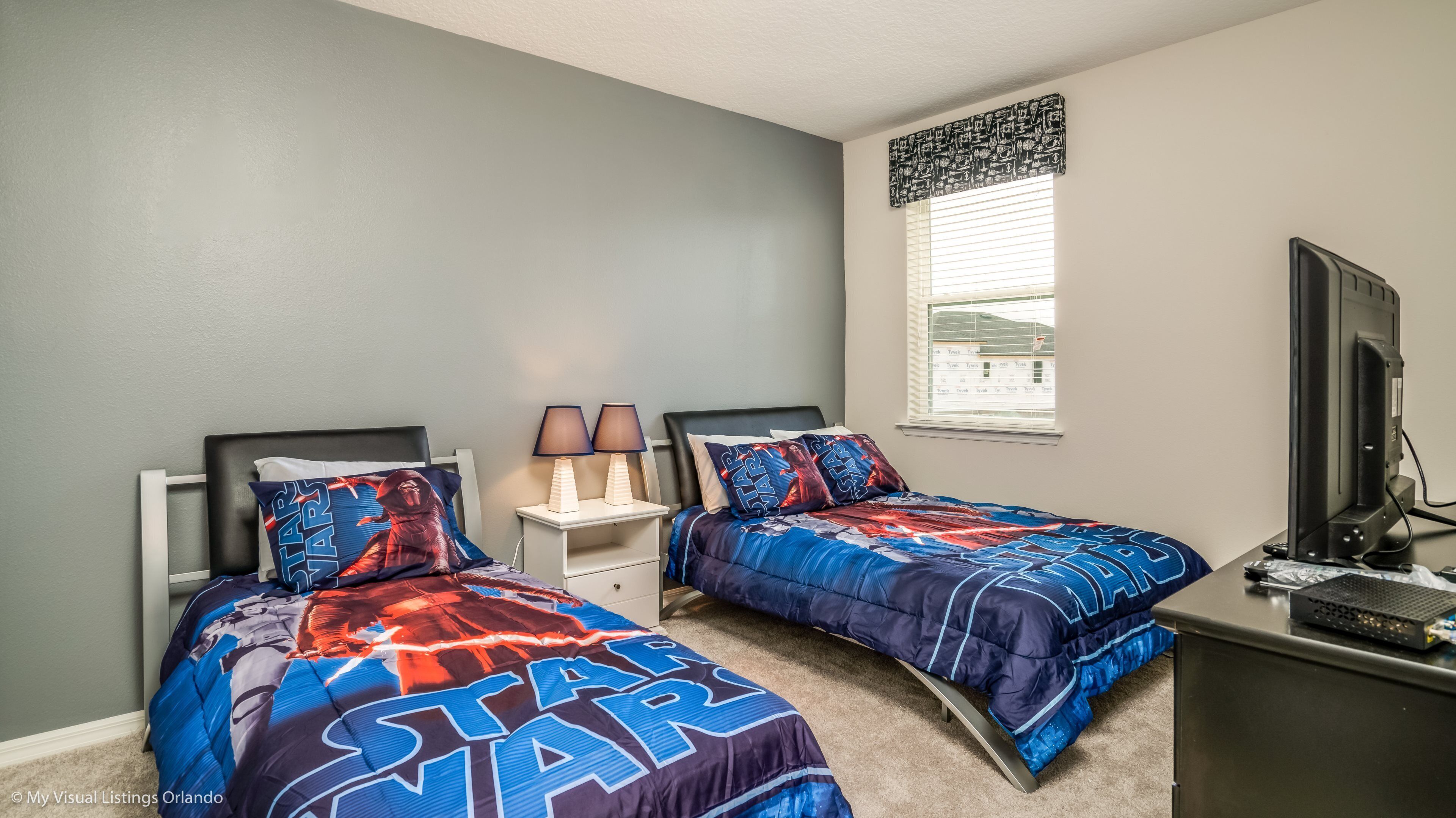 Quarto com duas camas, roupa de cama com tema Star Wars, mesa de cabeceira com luminárias, janela com persianas e uma TV em uma escrivaninha.