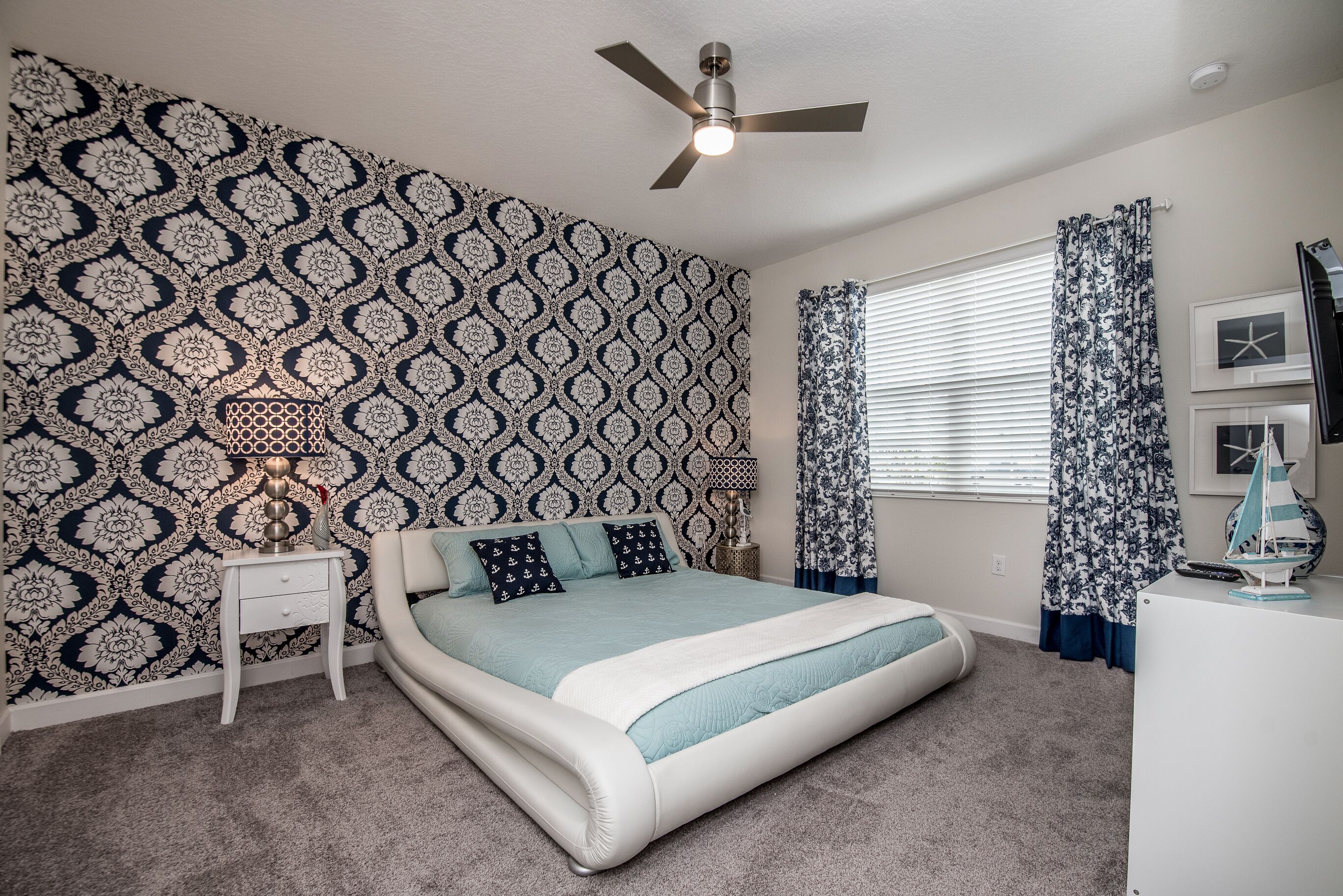 A bedroom with a patterned wallpaper, a bed with a blue and white bedspread, a ceiling fan, a nightstand with a lamp, and a window with curtains.