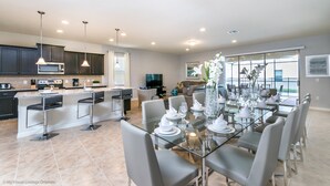 A modern kitchen with dark cabinets, a central island, and a dining area with a glass table and chairs. - Beautiful 8 Bed Villa Pool & Spa (Kissimmee)