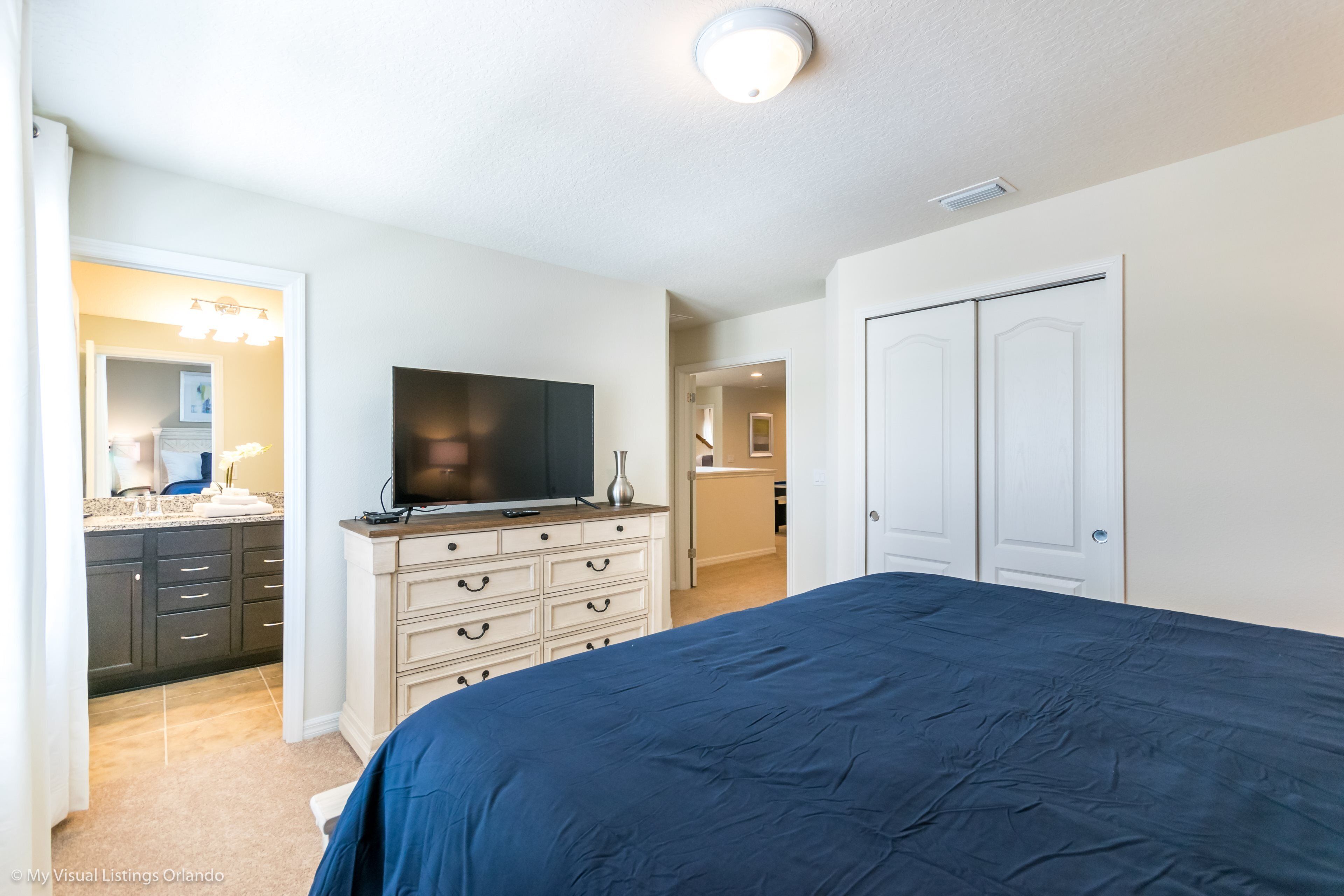 A bedroom with a bed, dresser, TV, and a door leading to another room.