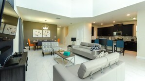 A spacious living area with a sofa, coffee table, and dining table. A kitchen with dark cabinets and a microwave is visible in the background.