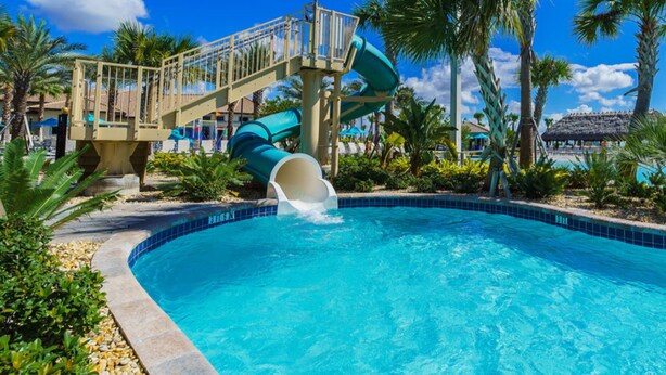 A swimming pool with a water slide, palm trees, and a clear blue sky.