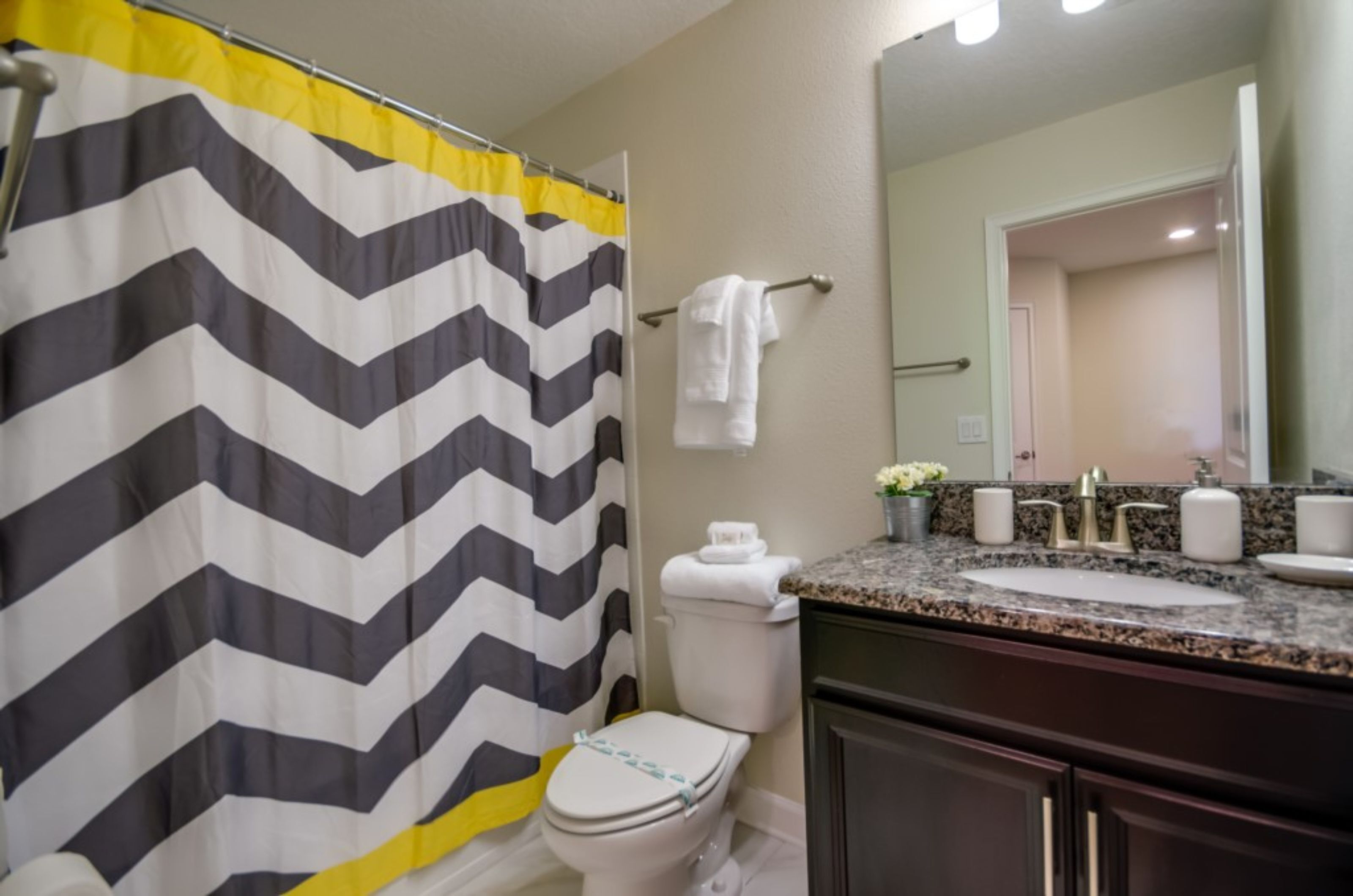 A bathroom with a black and white chevron shower curtain, a white toilet, a granite countertop sink, and a mirror above the sink.