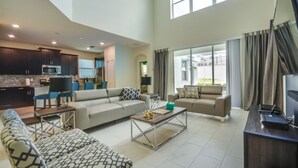 A spacious living area with a sofa, coffee table, and a TV stand. There is a kitchen area with dark cabinets and a microwave.