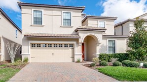 A two-story house with a beige facade, a tiled roof, and a brick driveway.