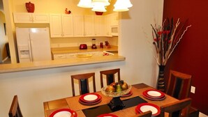 A dining area with a wooden table set for four, white cabinets, a red vase, and a vase with decorative branches. - Sun Lake IHR 3062 (Kissimmee)