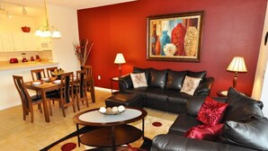 A living room with a black leather sofa, wooden dining table, and chairs. There is a painting on the wall and two lamps on side tables. - Sun Lake IHR 3062 (Kissimmee)