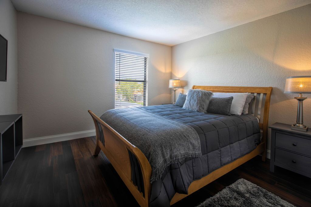 A bedroom with a wooden bed, two bedside lamps, a window with blinds, and a small bedside table.