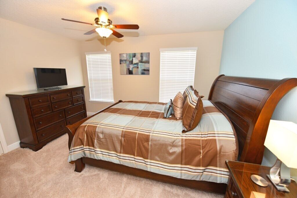 A bedroom with a bed, a dresser, a television, a ceiling fan, and two windows with blinds.
