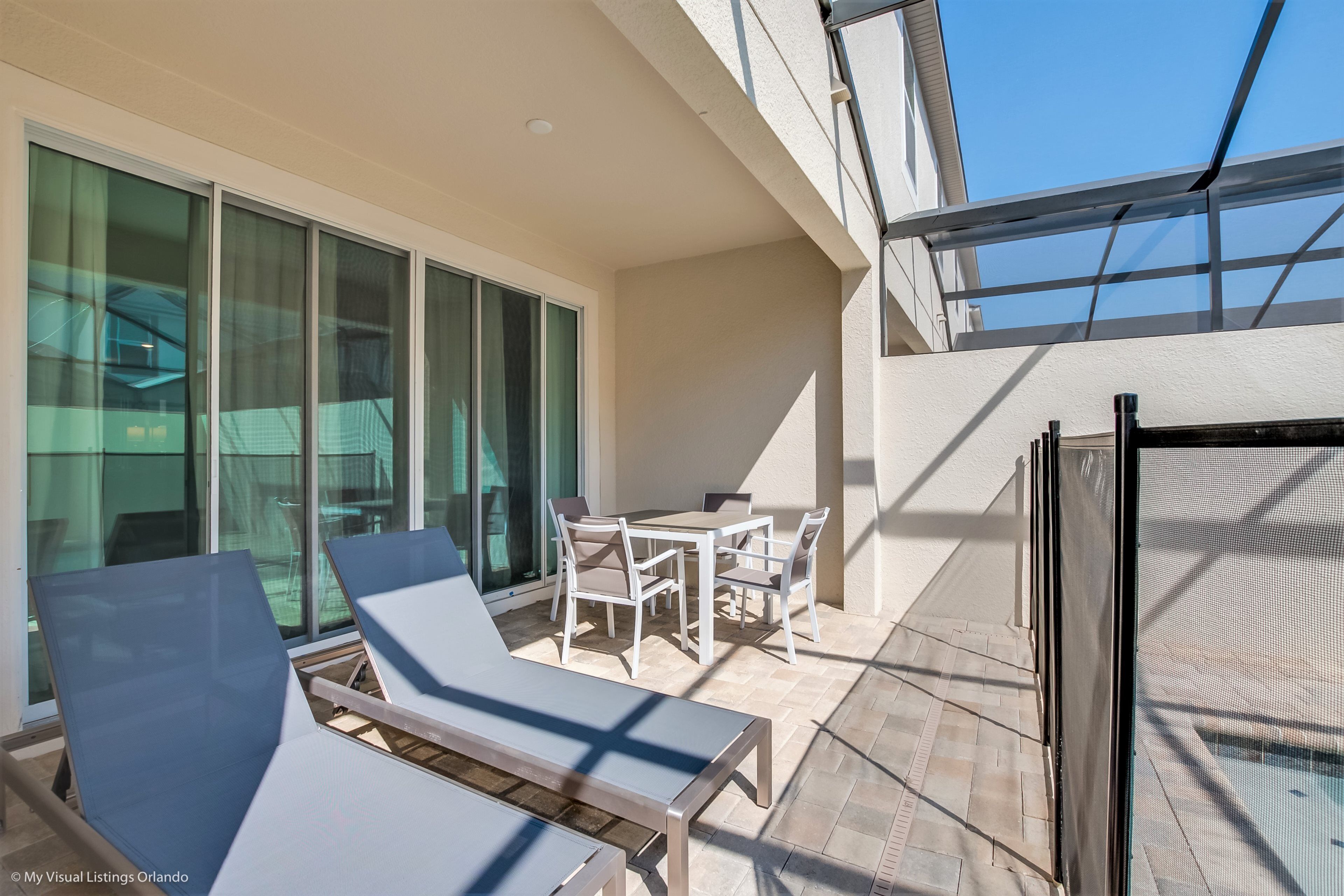 A sunlit outdoor patio with a table and chairs, a lounge chair, and a large window overlooking a pool.
