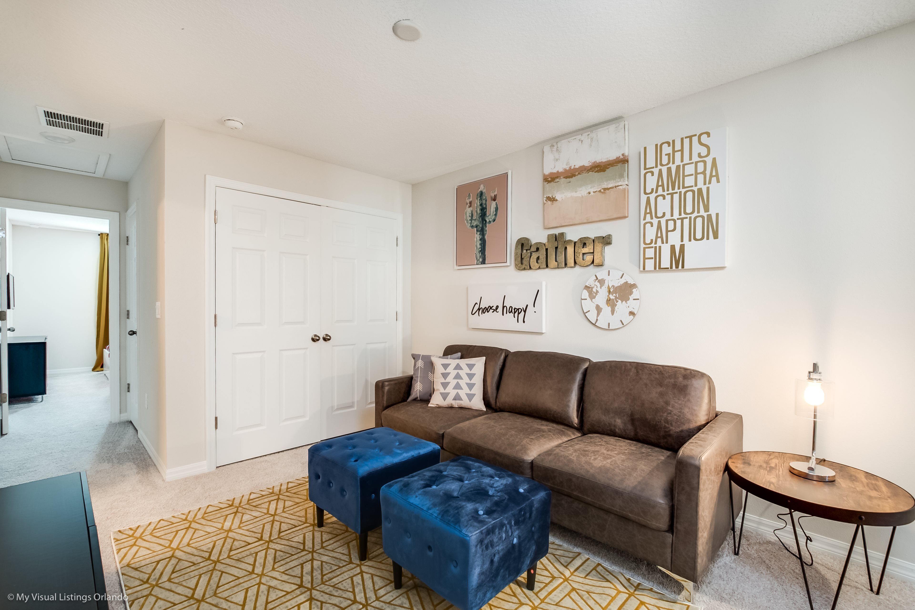 Un salon avec un canapé en cuir marron, deux poufs bleus, un tapis à motifs et des œuvres d’art murales, dont une représentation d’un cactus et un texte.