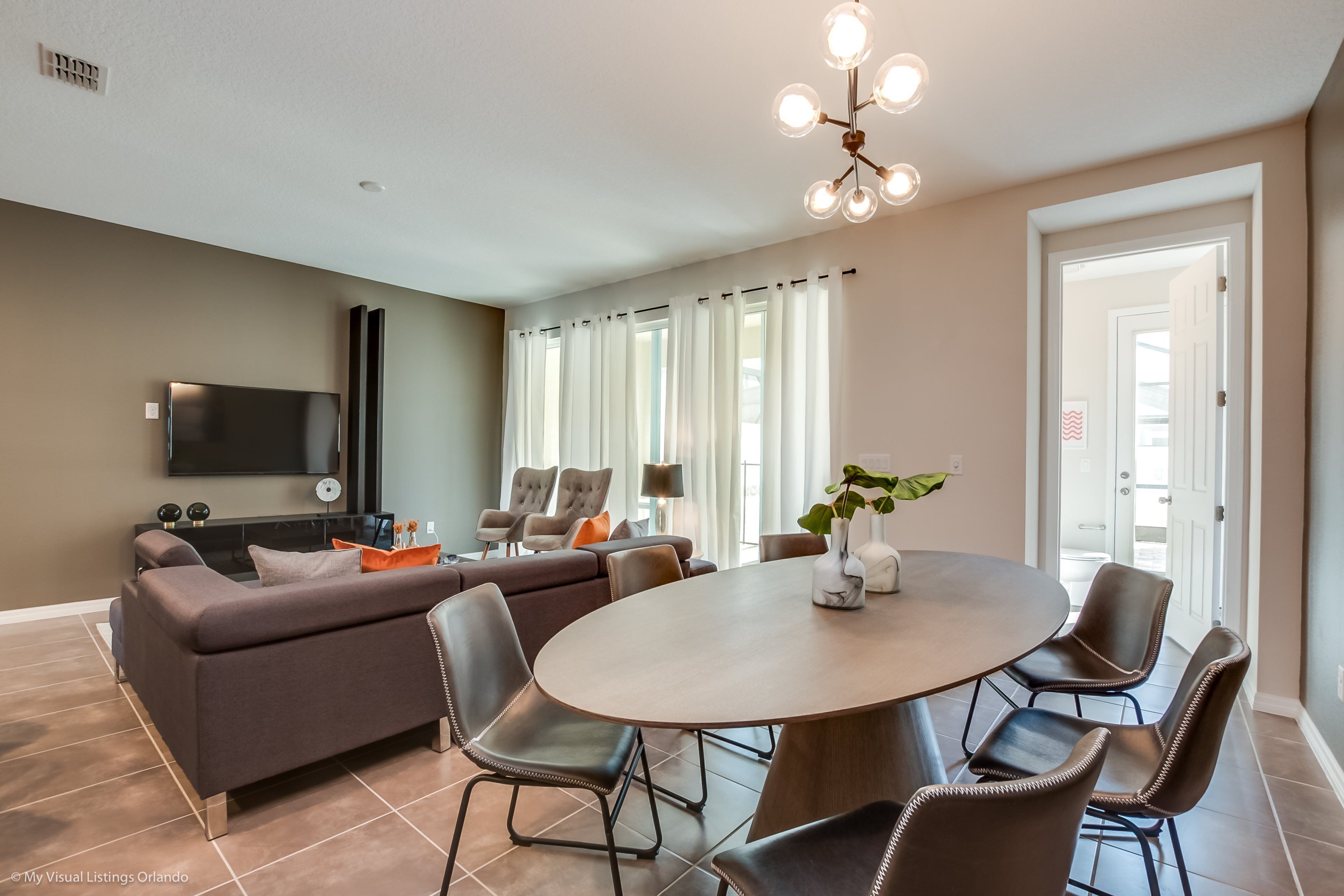 A modern living room with a large sofa, a dining table with chairs, a flat-screen TV mounted on the wall, and a chandelier.
