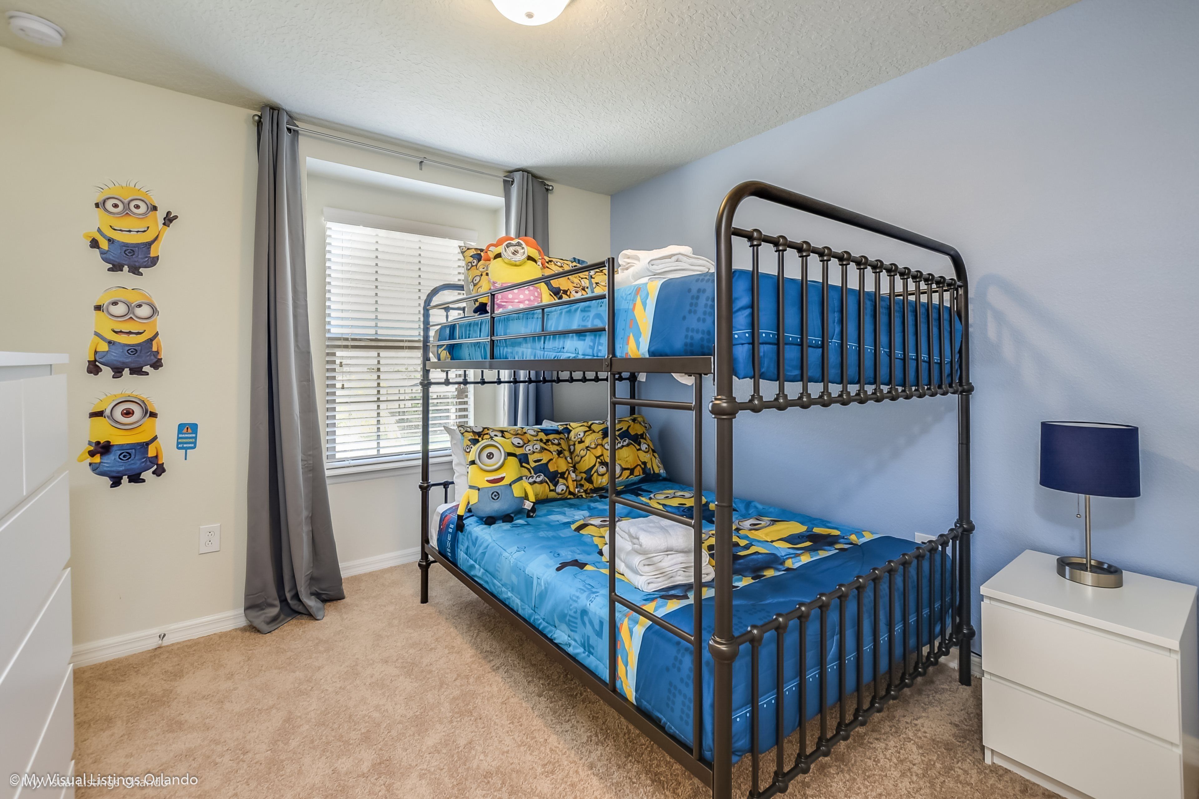 A bunk bed with a blue and yellow bedspread, a white nightstand with a lamp, and Minion wall decals.