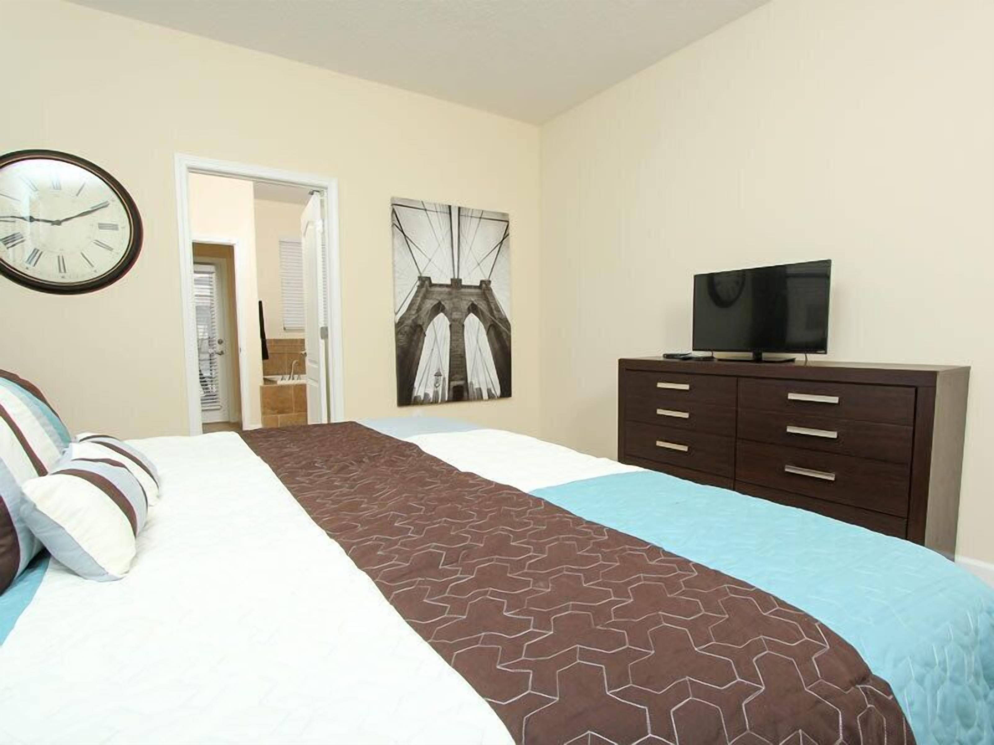 A hotel room with a bed, a TV on a dresser, a clock on the wall, and a door leading to a bathroom.