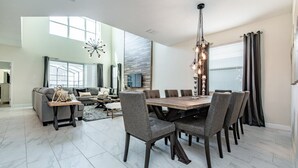 A spacious dining area with a large wooden table and multiple chairs, a modern chandelier, and a large window with curtains. - 5 Bedroom Resort Villa with Game Room & Pool (Kissimmee)