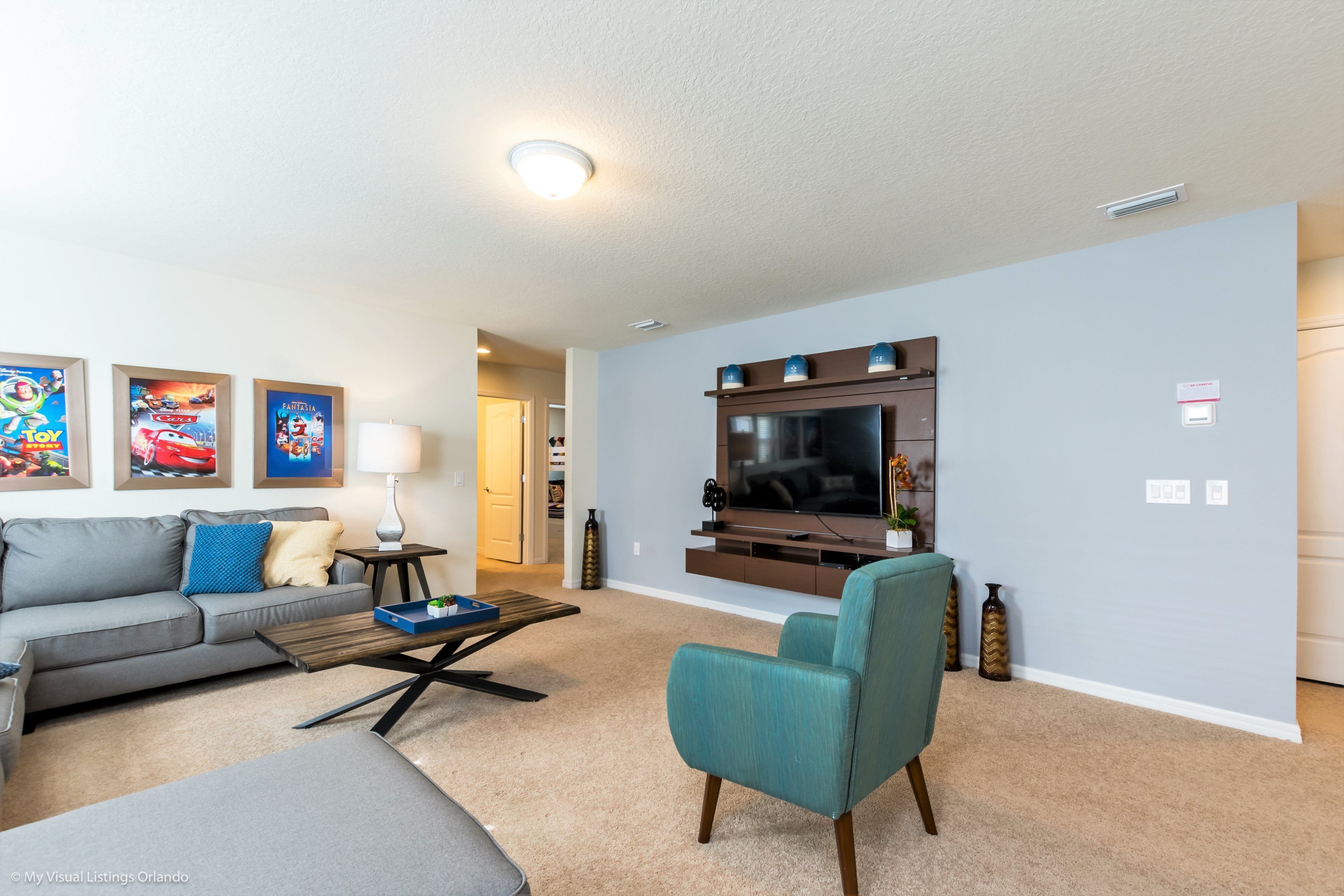 A living room with a grey sofa, teal armchair, and a flat-screen TV mounted on the wall.