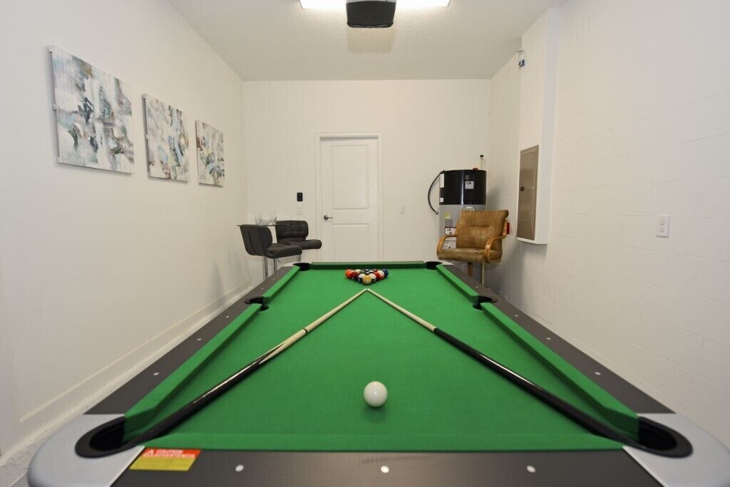 A pool table with a green felt surface, a white cue ball, and two pool cues. There are three abstract paintings on the wall.