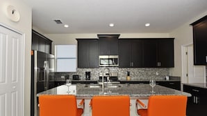 Una cocina moderna con armarios oscuros, un refrigerador de acero inoxidable, un microondas y una encimera de granito. Hay tres taburetes naranja alrededor de la isla.