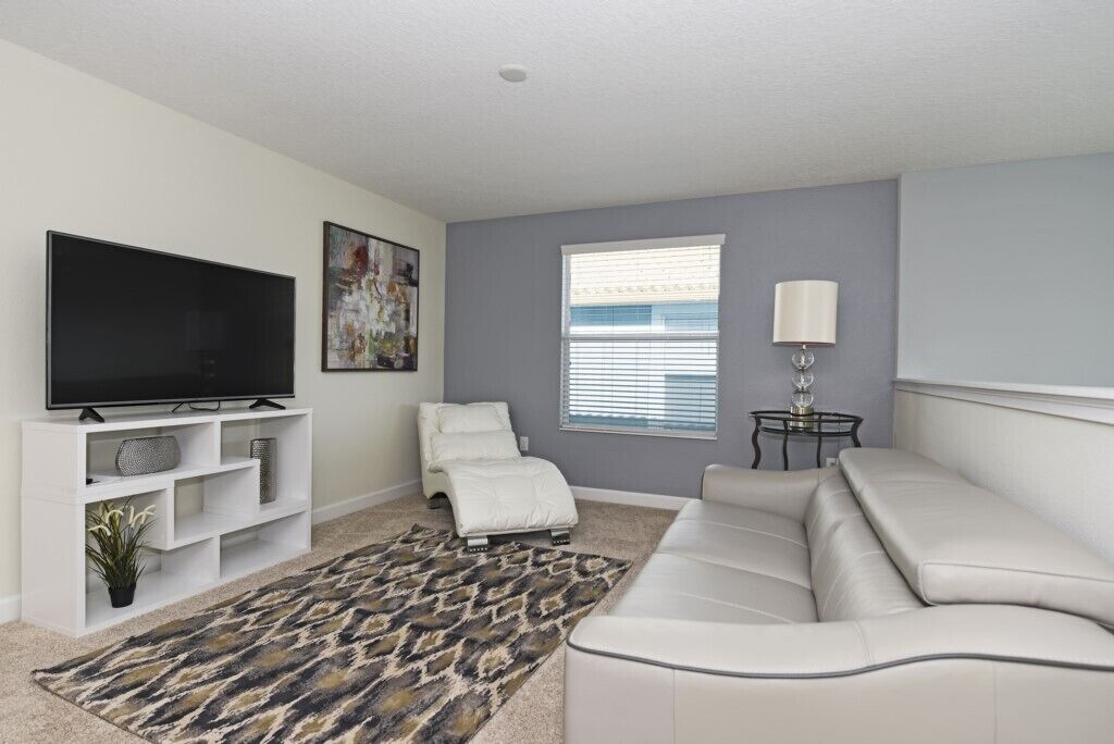 A modern living room with a flat-screen TV, a sofa, a recliner, a patterned rug, and a window with blinds.