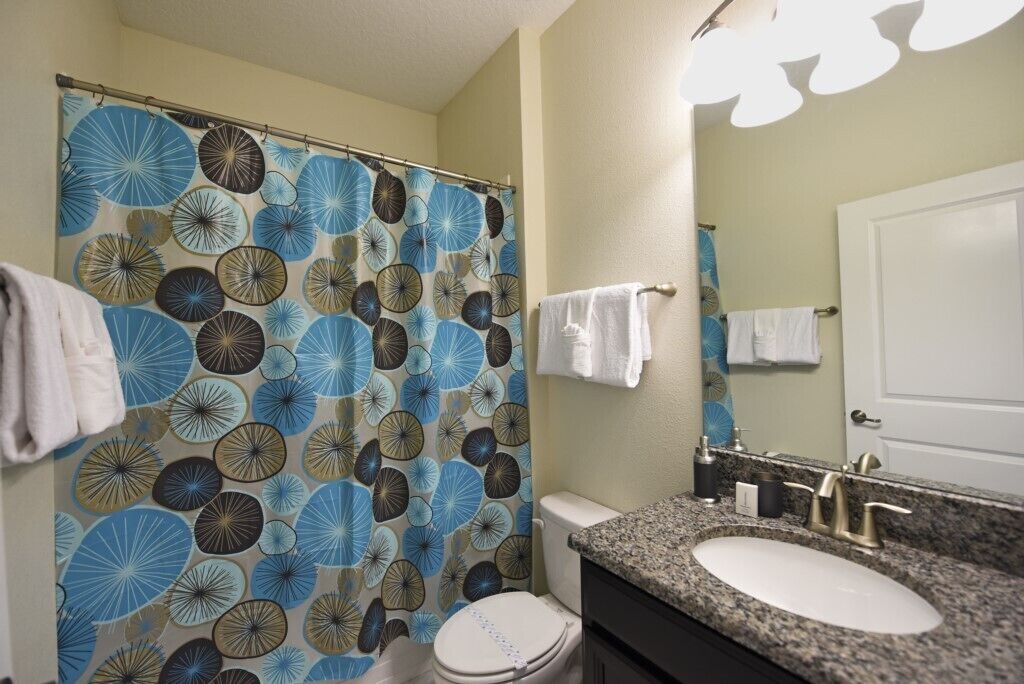 Ein Badezimmer mit Duschvorhang in blau-braunem Rundmuster, einer weißen Toilette, einem Waschbecken mit Granittischplatte und einem Spiegel über dem Waschbecken.