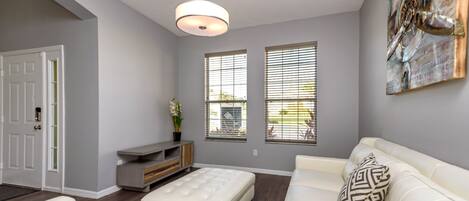 A modern living room with a white sofa, a matching ottoman, and a decorative pillow. There is a small side table with a plant, and a wall-mounted light fixture.