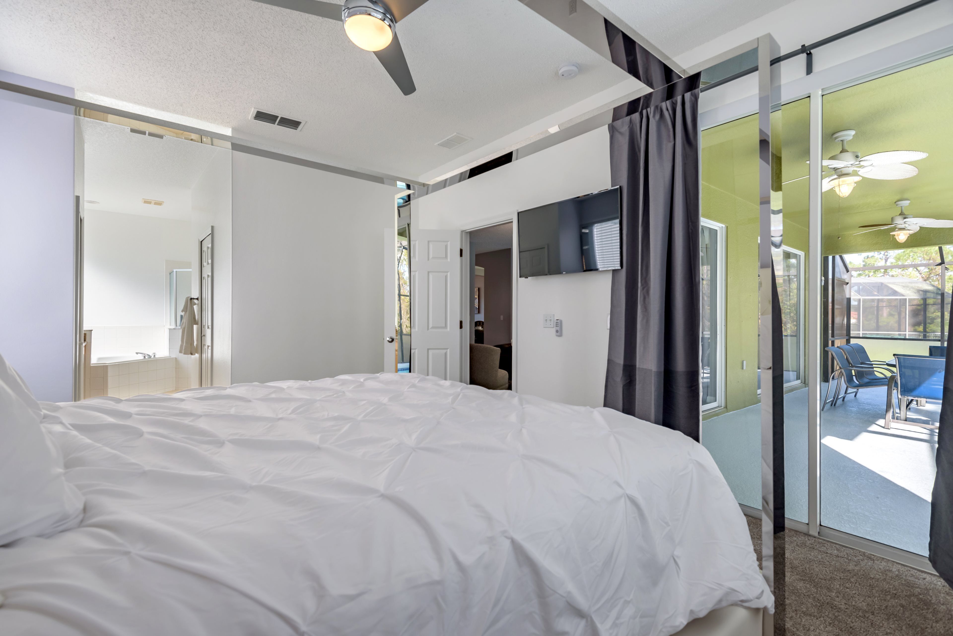 A bedroom with a large bed, a ceiling fan, a sliding glass door leading to a balcony with outdoor seating.
