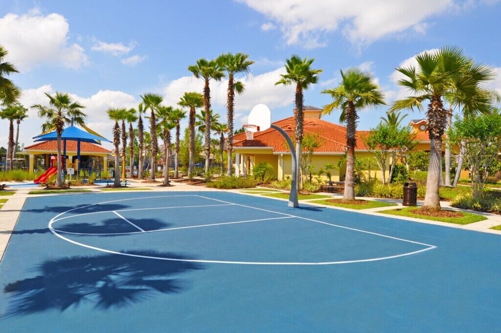 Una cancha de baloncesto rodeada de palmeras y un parque infantil con un tobogán.