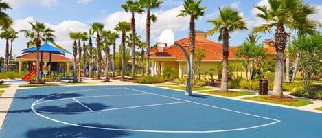 Una cancha de baloncesto rodeada de palmeras y un parque infantil con un tobogán.