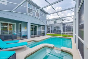 A modern indoor swimming pool with glass walls, a lounge area with blue cushions, and a view of the surrounding landscape.