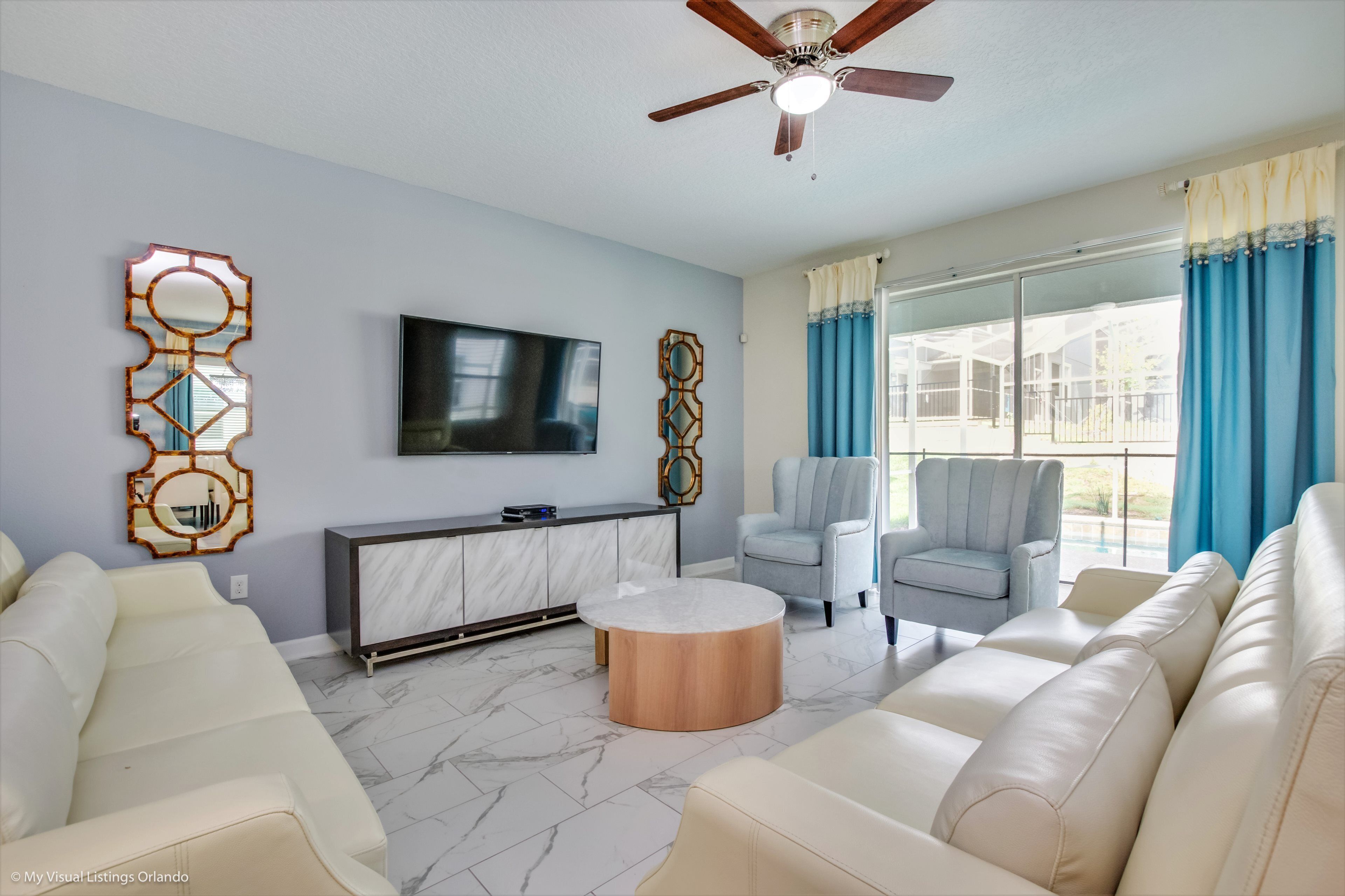 A modern living room with a marble floor, a ceiling fan, a flat-screen TV, a marble console, two armchairs, and a sofa.
