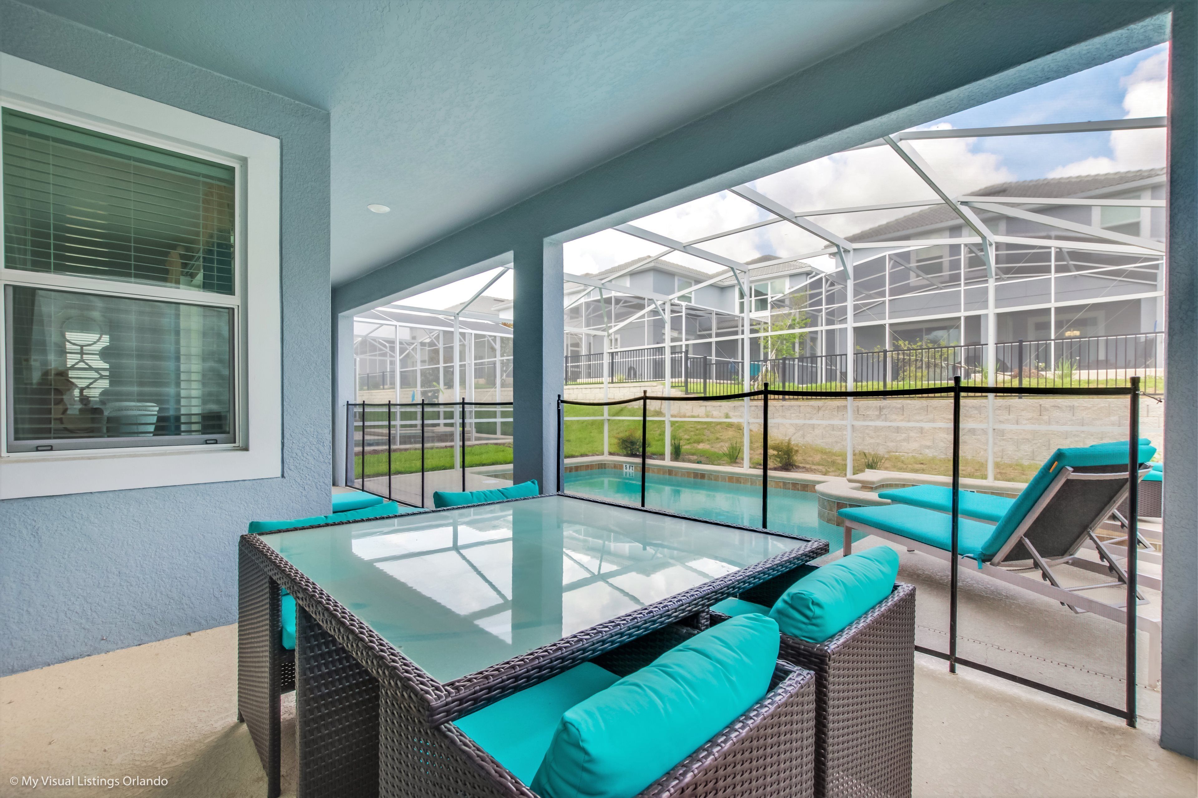 A covered patio with a glass table and wicker furniture, overlooking a pool and greenhouse.