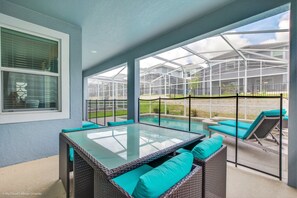 A covered patio with a glass table and wicker furniture, overlooking a pool and greenhouse.
