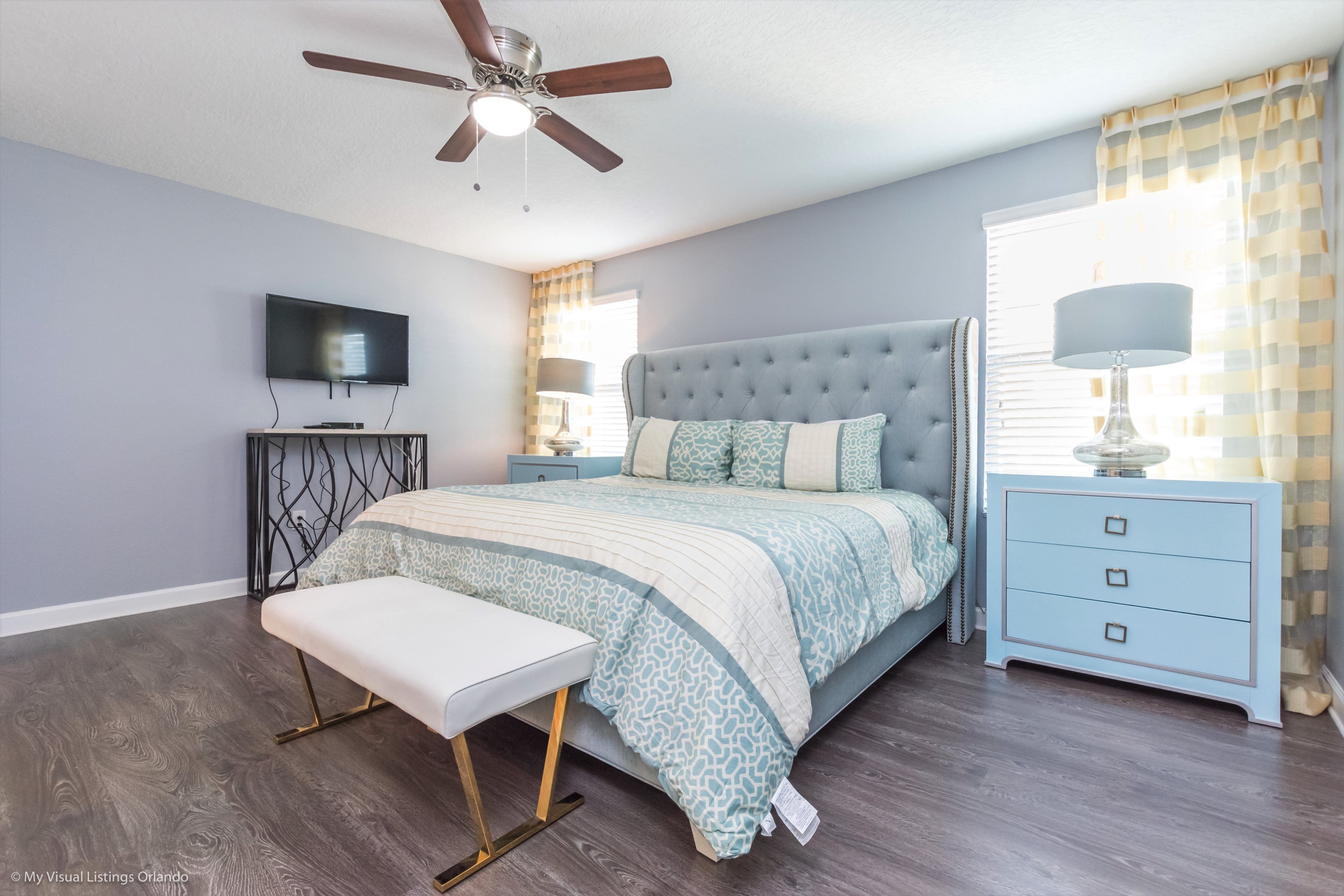 A bedroom with a tufted headboard bed, a bench at the foot of the bed, a TV mounted on the wall, a ceiling fan, and a window with curtains.