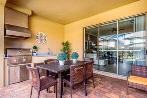 An outdoor patio with a grill, dining table, chairs, potted plants, and a view of a swimming pool through glass doors. - 2105mw Luxury 6 Bed Villa With Pool & Spa (Kissimmee)
