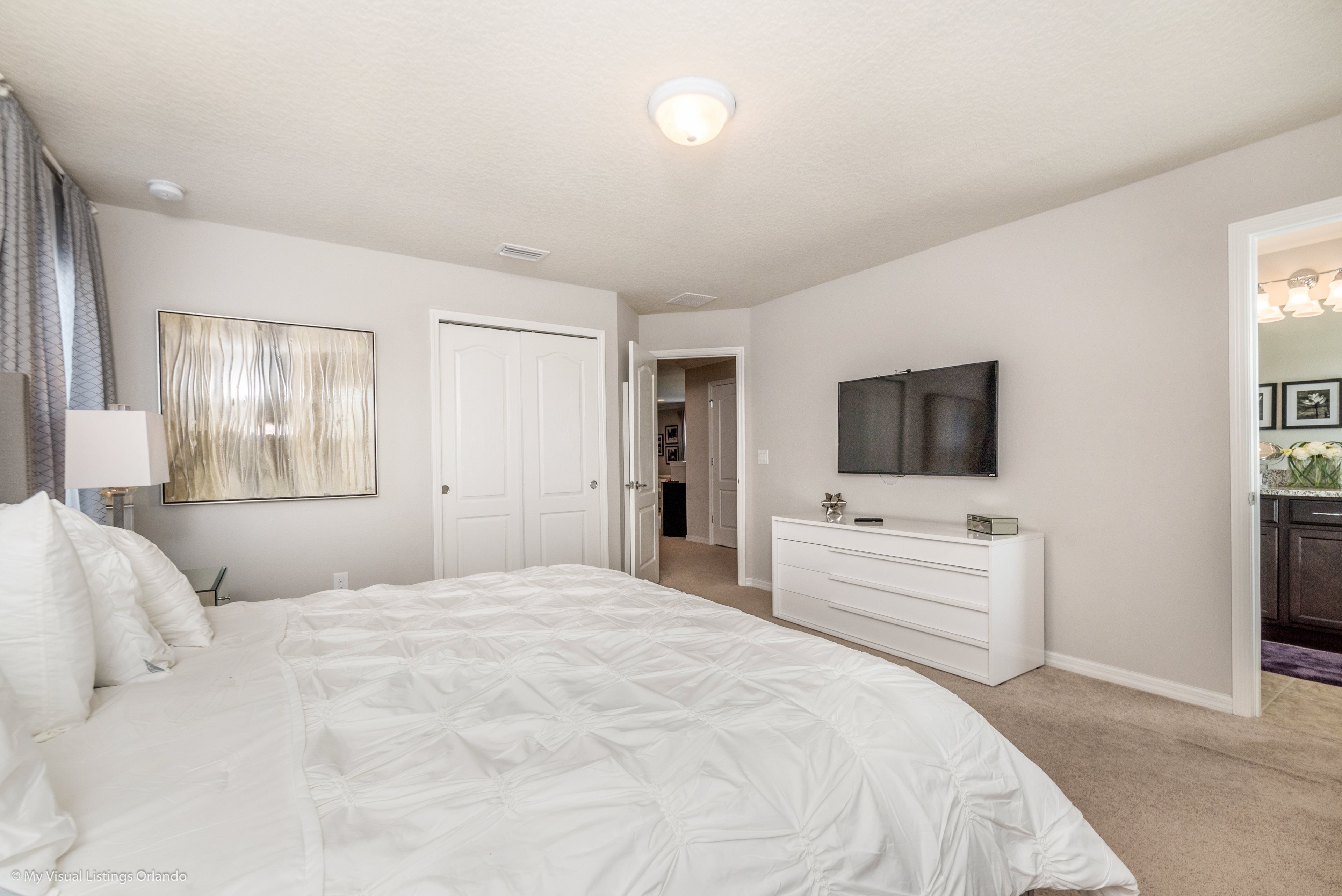 A bedroom with a large bed, a dresser, a flat-screen TV, and a door leading to another room.