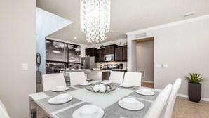 A modern dining area with a white table set for six, surrounded by white chairs. A decorative light fixture hangs from the ceiling, and a kitchen is visible in the background. - 2105mw Luxury 6 Bed Villa With Pool & Spa (Kissimmee)