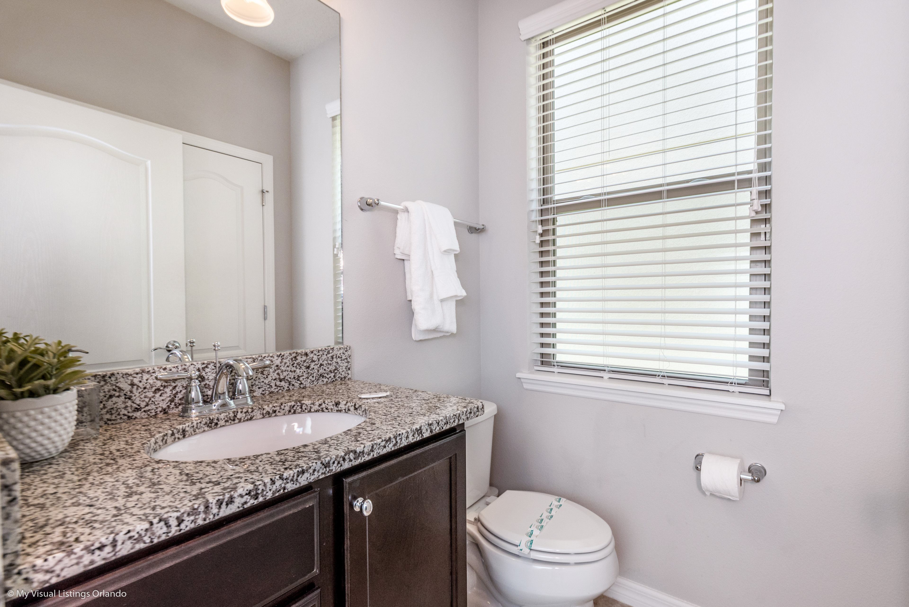 A bathroom with a granite countertop sink, a mirror, a towel rack, a toilet, and a window with blinds.