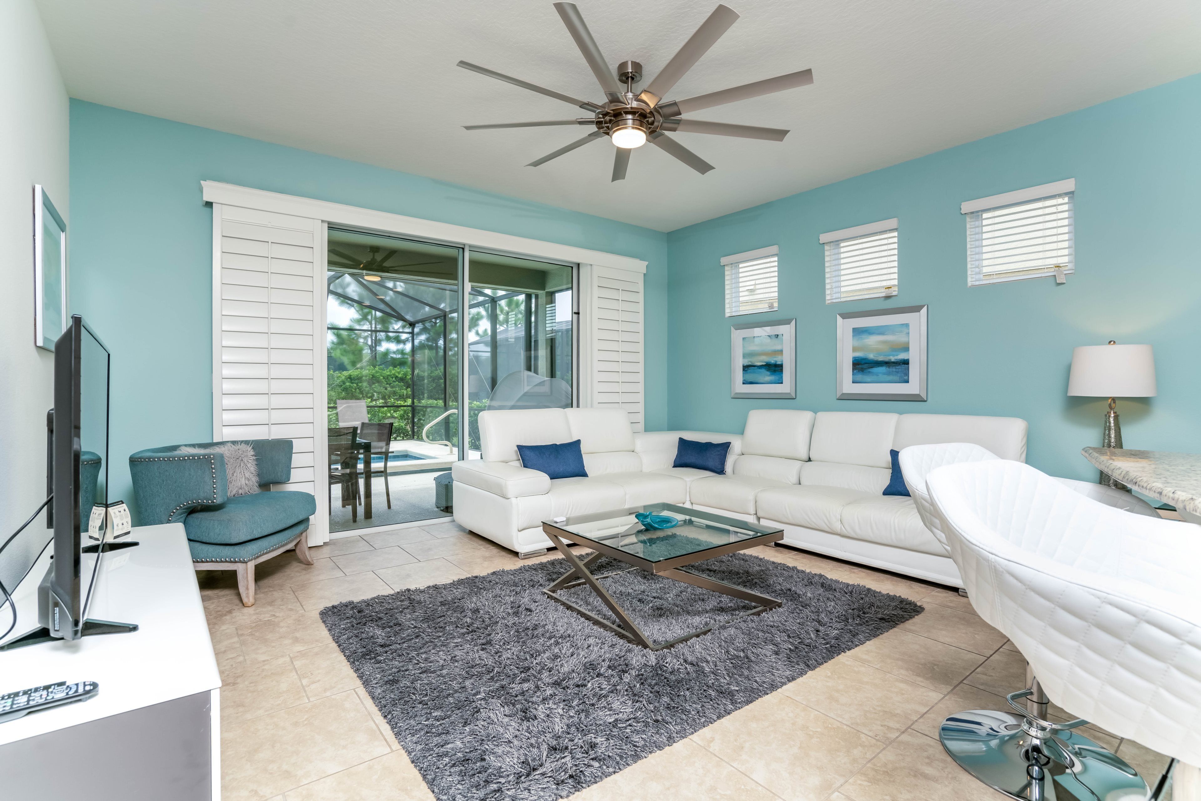 A modern living room with a white sofa, a glass coffee table, a ceiling fan, and a large window with blinds.