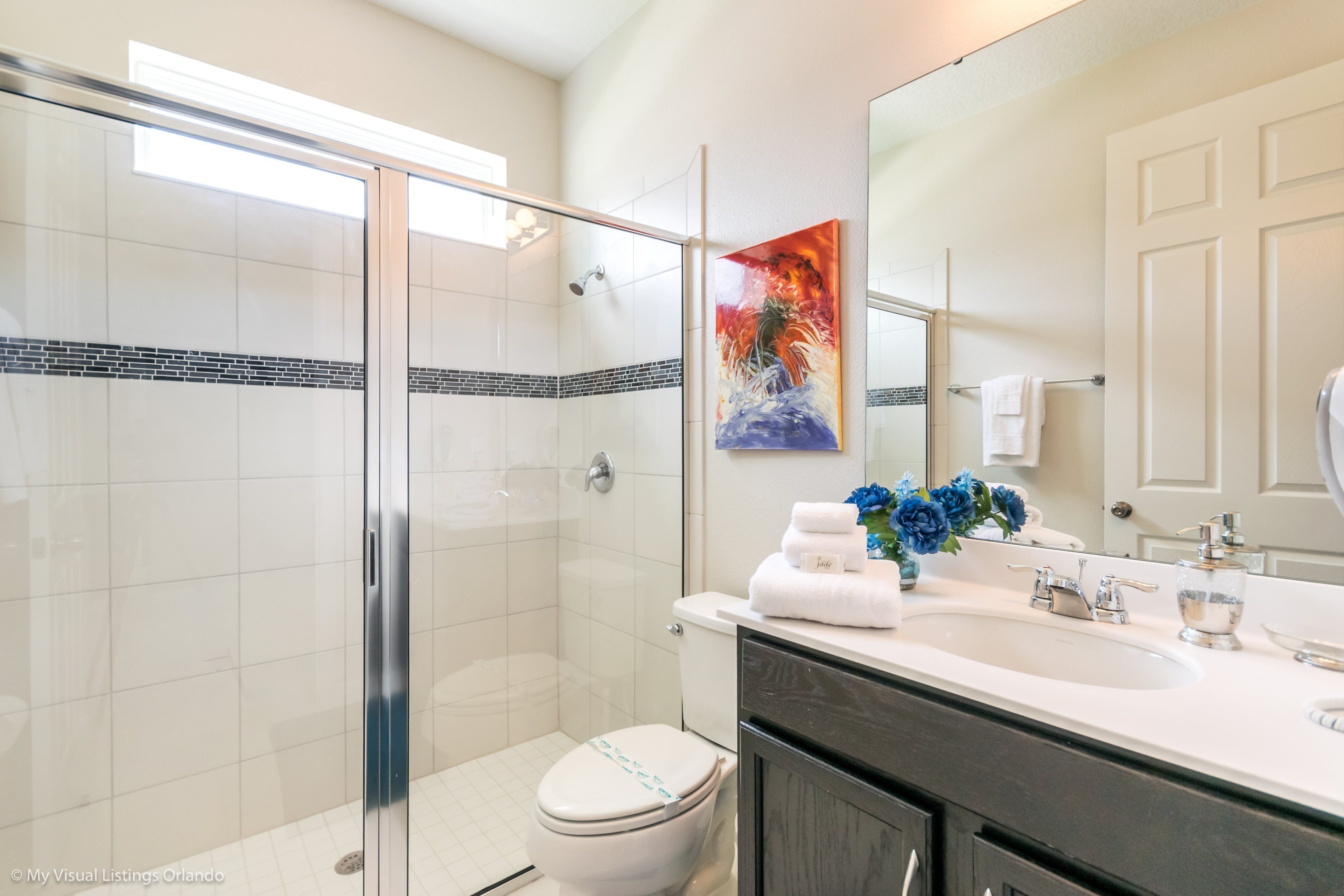 A modern bathroom with a glass shower enclosure, a white toilet, a sink with a mirror, and a decorative painting.