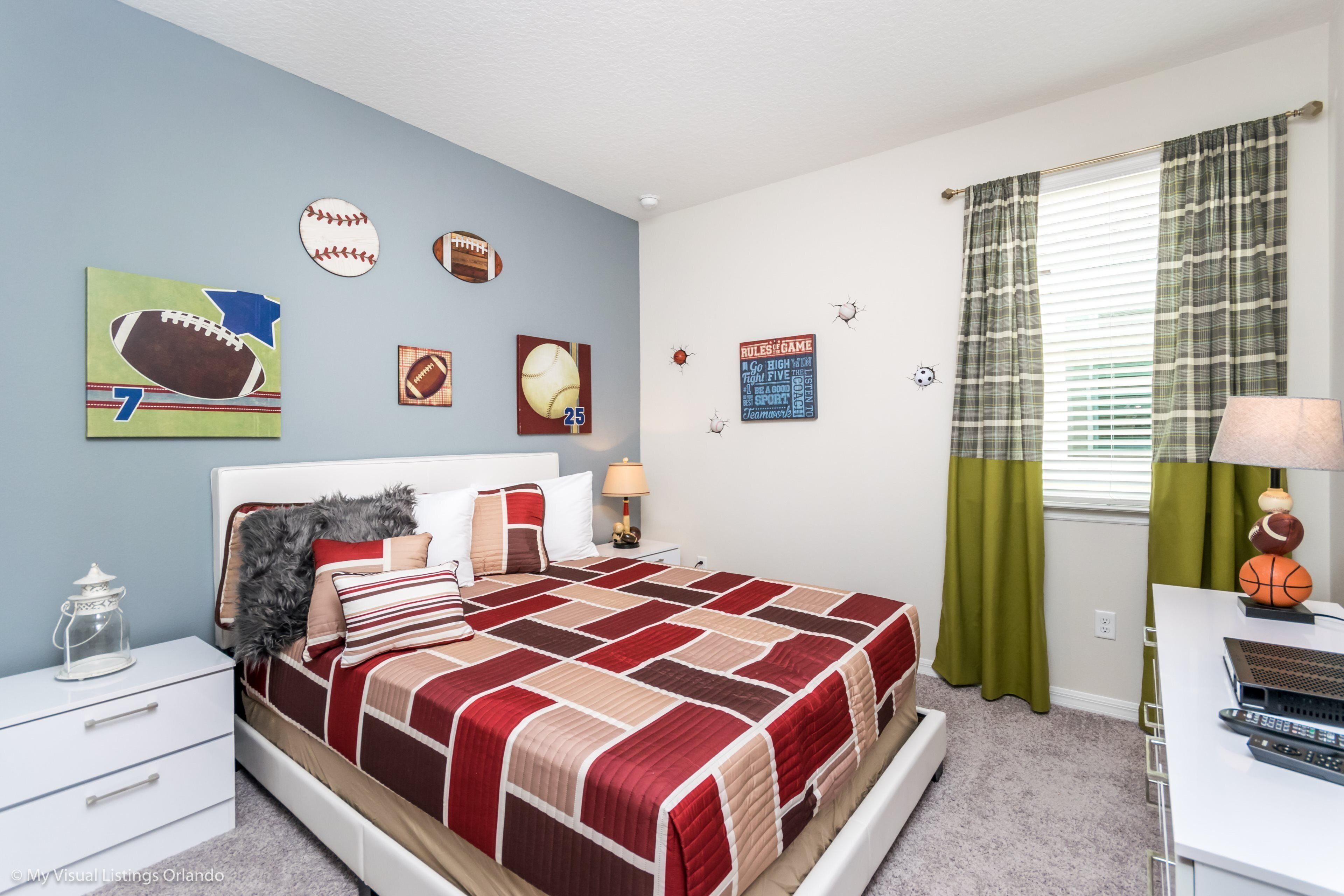A bedroom with a large bed, a nightstand, a lamp, a window with curtains, and sports-themed wall decorations.