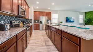 A modern kitchen with wooden cabinets, granite countertops, a stainless steel refrigerator, and a large island with a sink. - Solara Resort 9 Bedroom 6 Bathroom Resort Villa (Kissimmee)