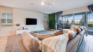 A modern living room with a large flat-screen TV, a sofa set, a glass coffee table, and a view of the outdoors through large windows. - Solara Resort 9 Bedroom 6 Bathroom Resort Villa (Kissimmee)