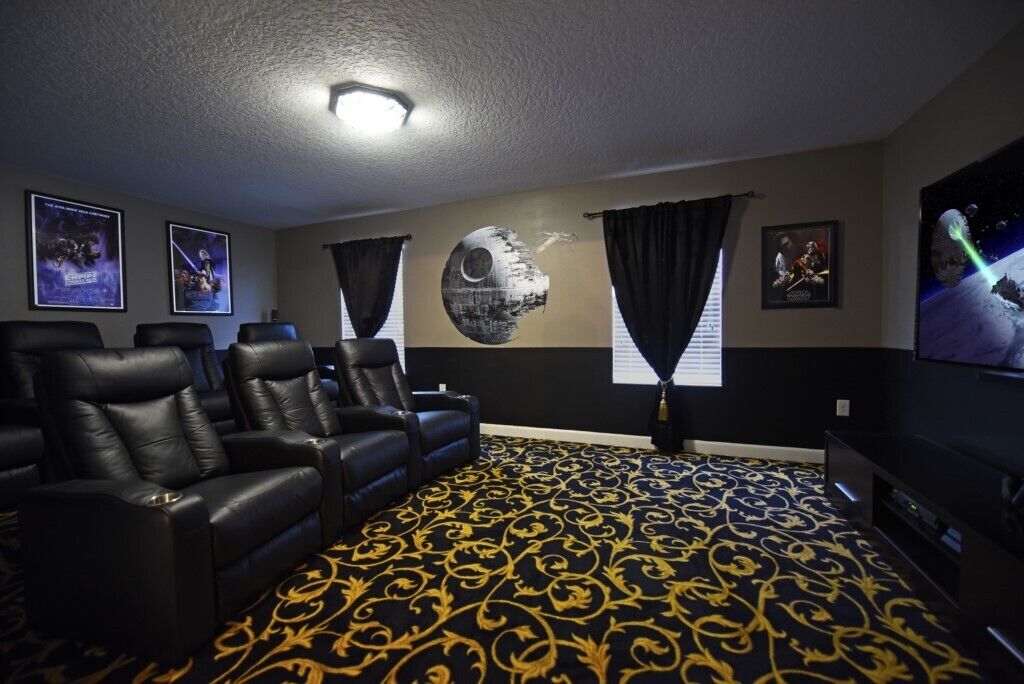 A home theater room with black leather recliners, a patterned carpet, Star Wars posters, and a large screen.