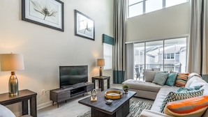 A modern living room with a flat-screen TV, a sofa, a coffee table, and a large window with curtains. - 5 Bdrm Champions Gate Luxury Villa (Davenport)
