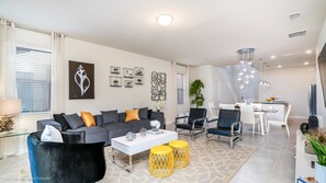 Un salon moderne avec un canapé gris, des poufs jaunes et une table basse blanche. On y trouve également un coin repas avec une table et des chaises blanches, et une cuisine en arrière-plan.