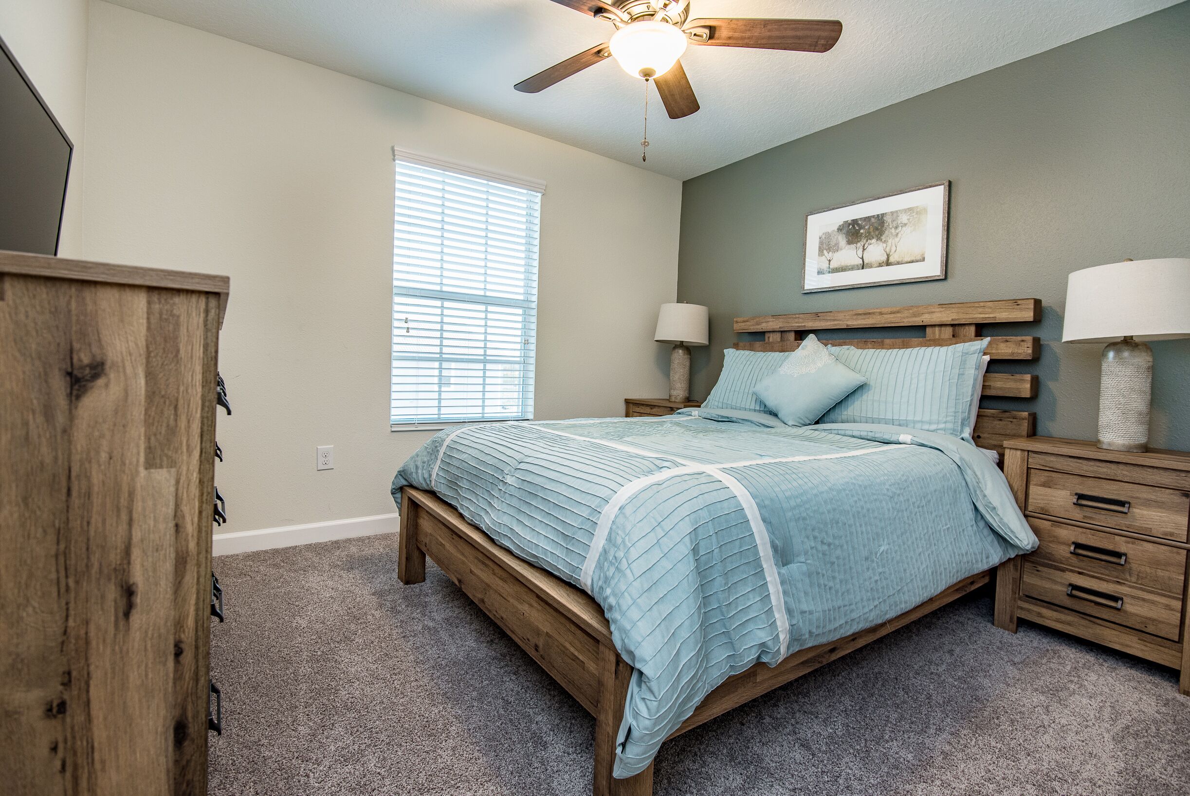 A bedroom with a wooden bed, two bedside tables, a ceiling fan, a window with blinds, and a framed picture on the wall.