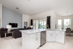 A modern kitchen with white cabinets, a dishwasher, and a large island. There is a dining area with a table and chairs, and a living room with a sofa and a TV.