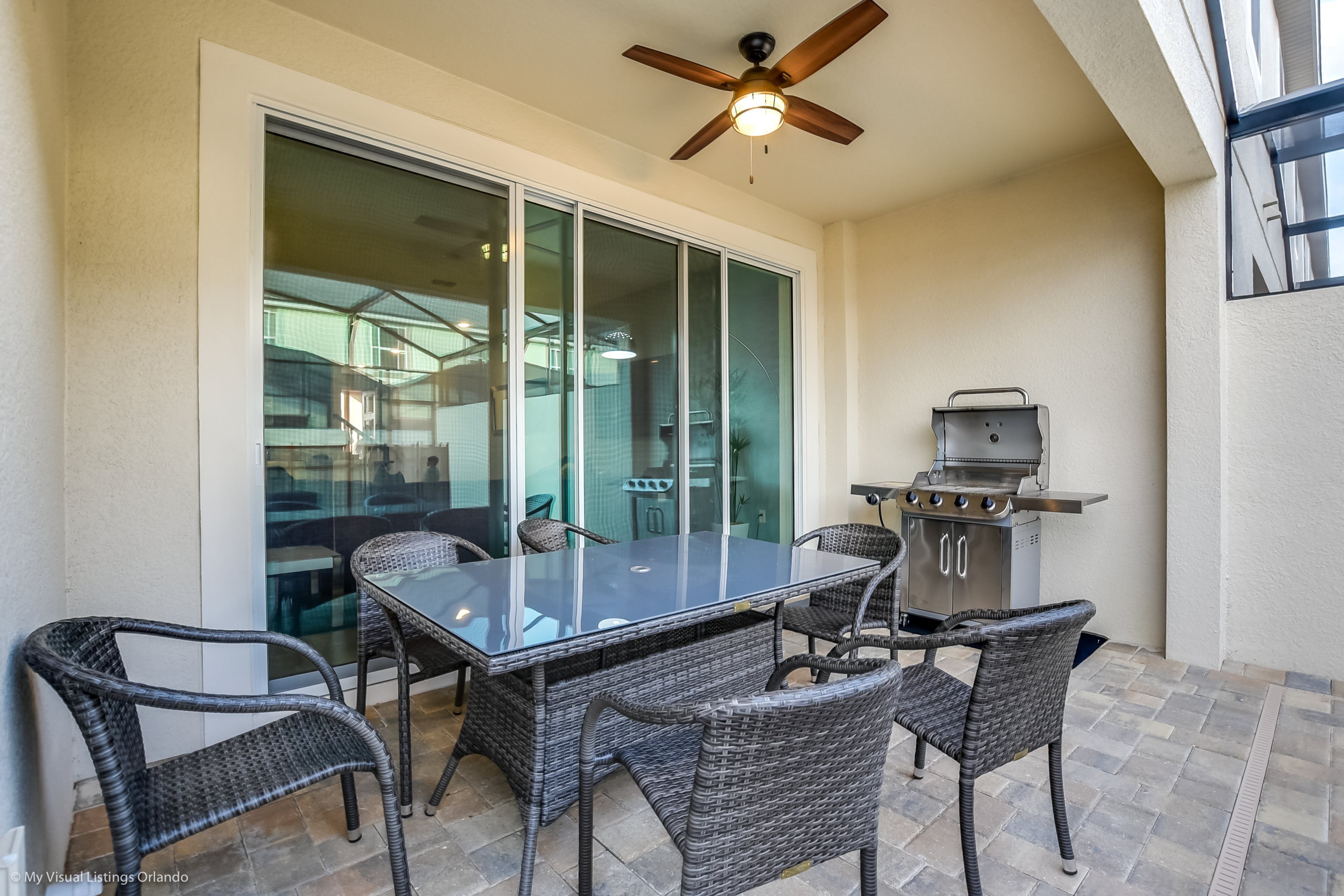 A patio with a glass-enclosed dining area, wicker chairs, a ceiling fan, and a barbecue grill.
