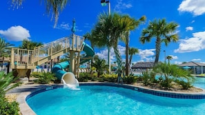Un espace piscine doté d’un toboggan aquatique, de palmiers et d’une structure à toit de chaume.