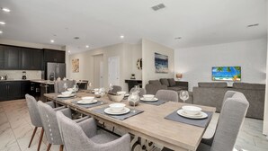A modern kitchen with dark cabinets, a dining area with a wooden table set for four, and a living area with a gray sofa and a TV displaying a beach scene.